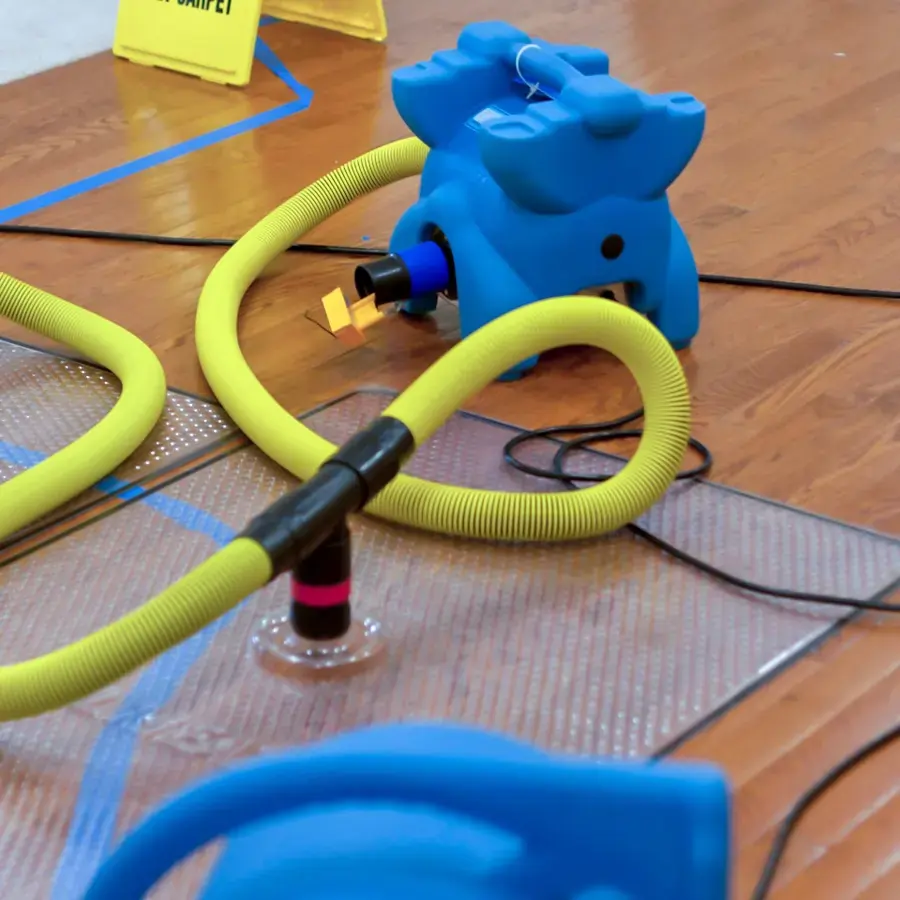 Blue air mover with yellow hoses running to a drying mat on a wood floor